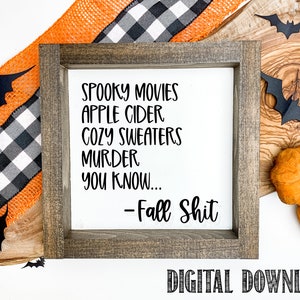 May include: A framed sign with a white background and black text that reads "Spooky Movies, Apple Cider, Cozy Sweaters, Murder, You Know... - Fall Shit". The sign is on a wooden surface with a black and white checkered ribbon and orange ribbon, and there are black paper bats and orange pumpkins on the surface.