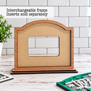 May include: A light brown wooden frame with an arched top and a rectangular center cutout. The frame sits on a tiered base. Text above the frame reads: Interchangeable frame Inserts sold separately. The frame is made of wood-grain material.