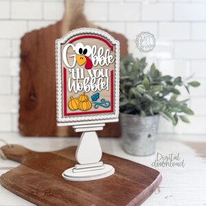 May include: A vertical wooden sign on a pedestal base featuring the text "Gobble 'til you wobble!" in white lettering. The design includes a turkey face, three orange pumpkins, a blue leaf, and blue swirls on a light wood background with a red border.