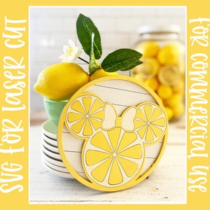 May include: A yellow wooden cutout of a Mickey Mouse head with lemon slices for ears. The cutout is on a white background with a wood grain texture. The cutout is surrounded by lemons and a glass jar of lemons.