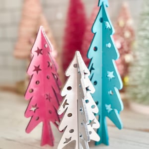 May include: Three wooden Christmas trees, one pink, one blue, and one white with silver glitter. Each tree has a star on top and cut-out stars on the sides.