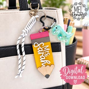 May include: A pencil-shaped keychain featuring a yellow body, a pink eraser, and a wooden tip. The name Mrs. Sarah is written in white script across the yellow section. It includes a white rope lanyard, a black heart-shaped carabiner, and a small translucent green dinosaur charm attached to a metal ring.