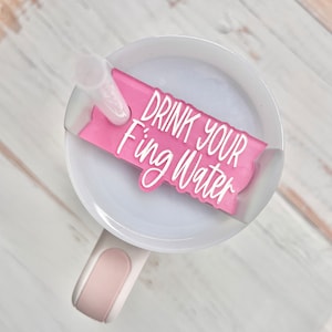 May include: A white tumbler with a pink handle and a clear straw. A pink acrylic tag with the words "DRINK YOUR Fing Water" in white lettering is attached to the lid. The tumbler is on a light wood surface.