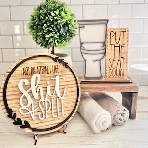 May include: A wooden sign with the text "Not an internet cafe' Shit & Split" in black and white lettering. The sign is round and has a black border. A small wooden stand with a toilet and a sign that says "Put the seat down" is in the background.