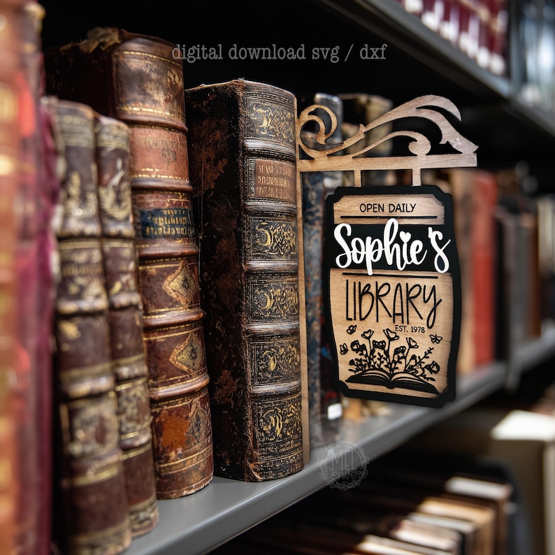 May include: A wooden sign for "Sophie's Library" with the text "Open Daily" and "Est. 1978". The sign is black and brown, with a floral design at the bottom. The sign is displayed on a shelf with antique books.