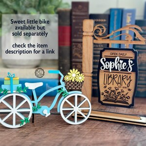 May include: A light blue and white decorative bicycle with a basket of yellow flowers, a gift, and a sign that reads "Sophie's Library" with the text "Open Daily". The sign is brown with a black border.