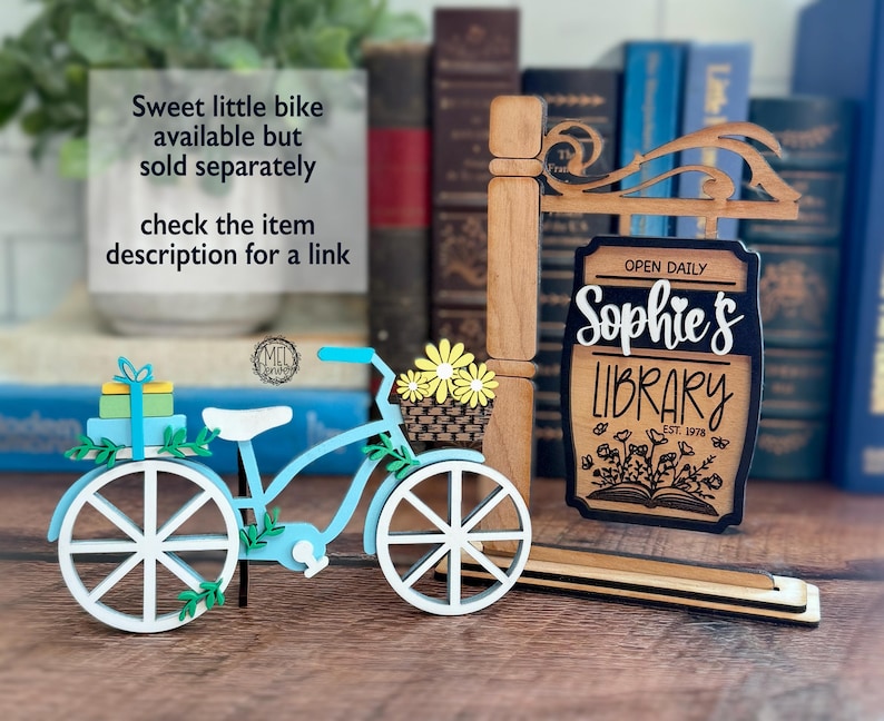 May include: A light blue and white decorative bicycle with a basket of yellow flowers, a gift, and a sign that reads "Sophie's Library" with the text "Open Daily". The sign is brown with a black border.