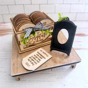 May include: A wooden box containing multiple engraved cards labelled Scripture of the Week, decorated with a black and white gingham ribbon and green leaves. A black arched display stand with an oval cutout sits next to a single wooden card.