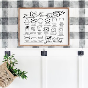 May include: A white sign with black text and symbols that reads "The Laundry Room". The sign includes a variety of laundry care symbols, such as bleach, do not bleach, dry clean, do not dry clean, machine wash normal, machine wash gentle, hand wash, machine wash cold, machine wash hot, do not tumble dry, tumble dry low heat, tumble dry gentle, do not iron, iron low heat, iron high heat. The sign also includes the text "Check your pockets".