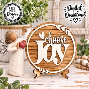 May include: A round wooden sign featuring the text "choose JOY" in white lettering, accented by a white heart and leaf border. Beside the sign stands a carved figurine of a figure in a long textured robe holding a small red heart.