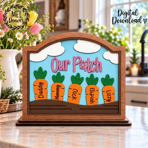 May include: A layered wooden sign featuring a light blue sky with white clouds and a brown soil base. The text "Our Patch" is in pink, and five orange carrots with green tops are labeled with the names Daddy, Mommy, Nick, Elishiah, and Lizzy.