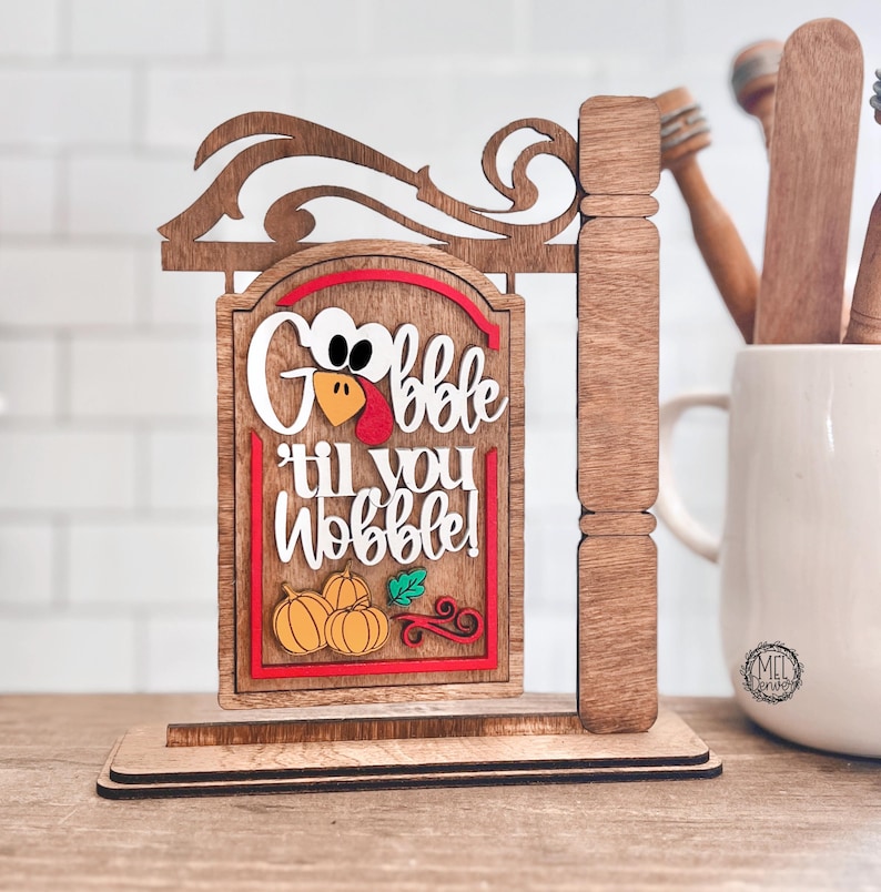 May include: A wooden tabletop sign featuring a hanging rectangular plaque with the text "Gobble 'til you Wobble!" in white lettering. The design includes a turkey face, orange pumpkins, a green leaf, and red swirls, all mounted on a brown base.