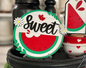 May include: A round wooden sign featuring the word "sweet" in black script, two small red hearts, a red watermelon slice with black seeds, and white daisy accents. Beside it hangs a rectangular wooden tag with a red watermelon slice graphic.