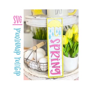May include: A pink and yellow wooden sign with the word "Spring" in pink letters and a yellow umbrella with blue raindrops. The sign is on a white tiered tray with a white mug, a basket of white eggs, and a potted plant with yellow flowers.