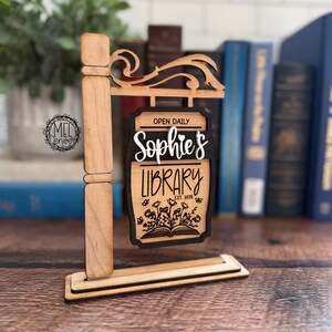 May include: Wooden signpost with the words "Sophie's Library" in a decorative font. The sign also includes the text "Open Daily" and "Est. 1978". The sign features a floral design and is made of light brown wood with black accents.