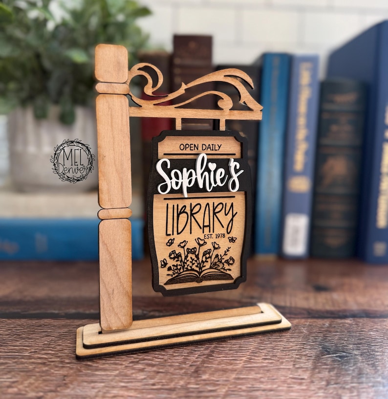May include: Wooden signpost with the words "Sophie's Library" in a decorative font. The sign also includes the text "Open Daily" and "Est. 1978". The sign features a floral design and is made of light brown wood with black accents.