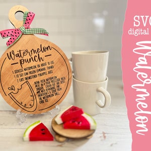 Op de afbeelding: Een houten snijplank met een handvat en een gegraveerd recept voor watermeloenpunch. Het recept bevat 1 zaadloze watermeloen, 1 blik bevroren limonade en 2 kopjes ongezoete ananassap. De plank ligt op een wit oppervlak met een kleine houten kom en watermeloenplakjes.