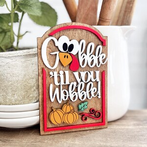 May include: A rectangular wooden sign with a rounded top featuring the text "Gobble til you Wobble!" in white 3D lettering. The sign includes a turkey face icon, three orange pumpkins, a green leaf, and red decorative swirls on a wood grain background.