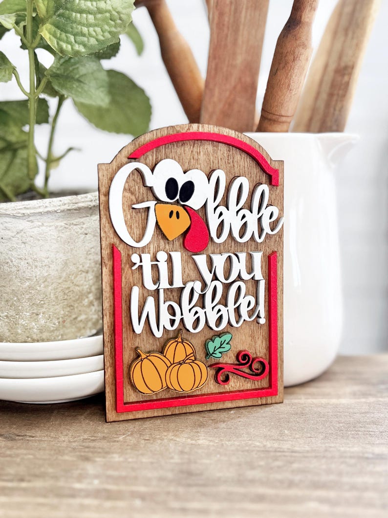 May include: A rectangular wooden sign with a rounded top featuring the text "Gobble til you Wobble!" in white 3D lettering. The sign includes a turkey face icon, three orange pumpkins, a green leaf, and red decorative swirls on a wood grain background.