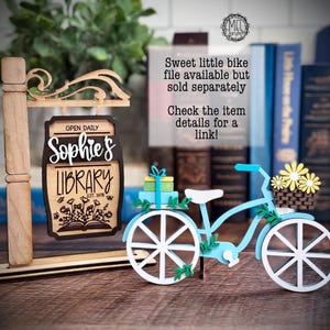 May include: A wooden sign for "Sophie's Library" with a light blue bicycle decoration. The sign is brown with white lettering and a floral design. The bicycle has white wheels, a basket of yellow flowers, and a small gift.