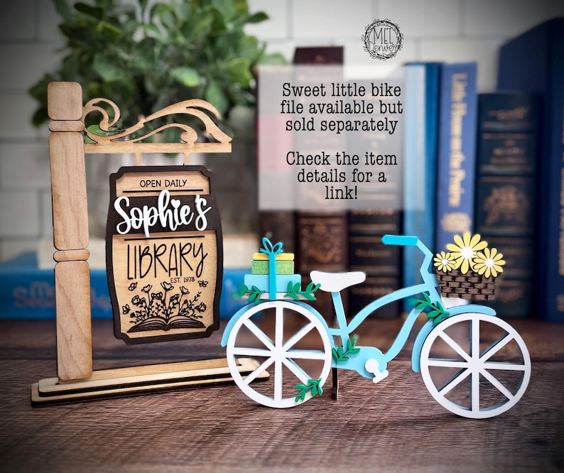 May include: A wooden sign for "Sophie's Library" with a light blue bicycle decoration. The sign is brown with white lettering and a floral design. The bicycle has white wheels, a basket of yellow flowers, and a small gift.