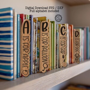 Può includere: Segnalibri alfabetici in legno con scritte nere, che compongono "A is for Apple", "B is for Balloon", "C is for Car" e "D is for Dog". I segnalibri sono posizionati tra una fila di libri colorati su uno scaffale. Il testo in alto recita "Digital Download SVG / DXF Full alphabet included".