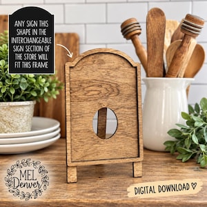 May include: A wooden sign frame with an arched top and a circular cutout in the center. The frame features a natural wood grain finish and stands on two small feet. Text on the image states: ANY SIGN THIS SHAPE IN THE INTERCHANGEABLE SIGN SECTION OF THE STORE WILL FIT THIS FRAME.