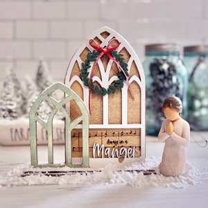 May include: A wooden Christmas church decoration with a green wreath and red bow, a green stained glass window, and the text "Away in a Manger". A small white figurine of a person praying is in the foreground.