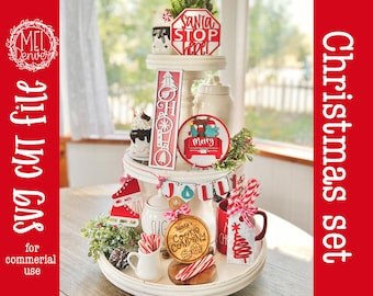 May include: A white three-tiered wooden tray decorated with Christmas-themed items including a Santa Stop Here sign, a Merry truck ornament, a Jolly banner, a Santa's Cookie Company wooden round, red ice skates, and a red and white striped candy cane.