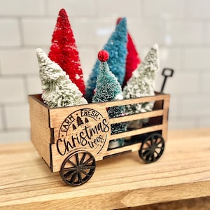 May include: A wooden wagon filled with miniature Christmas trees. The wagon is decorated with the words "Farm Fresh Christmas Trees".