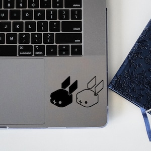 Peut inclure: Un ordinateur portable argenté avec un clavier noir et un autocollant de lapin noir et blanc sur le pavé tactile. Un carnet bleu foncé à motif floral et un stylo argenté se trouvent à droite de l'ordinateur portable.