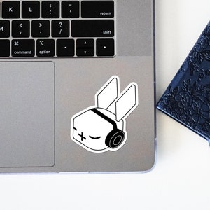 May include: A white sticker of a cartoon character with large ears, a plus sign on its face, and headphones. The sticker is on a silver laptop.