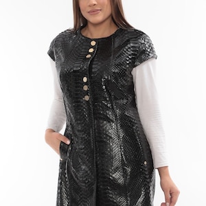 Pode incluir: Um casaco preto sem mangas, com comprimento até ao joelho, com um padrão de pele de cobra. O casaco apresenta botões dourados e um bolso. A modelo está a usar uma camisa cinzenta clara de manga comprida por baixo.