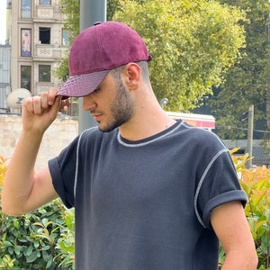 Peut inclure: Casquette de baseball bordeaux avec une visière texturée marron foncé. Le chapeau est porté avec un t-shirt noir à manches courtes avec des coutures blanches autour de l'encolure et des manches. Une montre-bracelet argentée est visible.