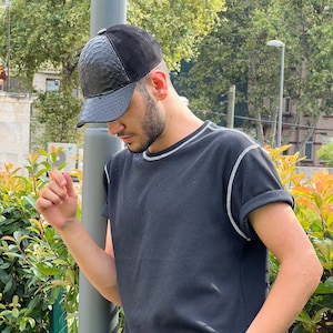 Może przedstawiać: Czarna czapka z daszkiem z fakturą imitującą skórę węża i zamszową górą. Czapka jest połączona z czarną koszulką z białymi szwami i podwiniętymi rękawami. Widoczny jest srebrny zegarek z zieloną tarczą.