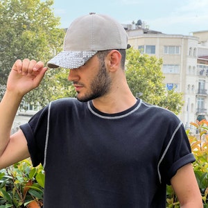 Może przedstawiać: Beżowa czapka z daszkiem z daszkiem wężowym. Osoba nosi czarny t-shirt z białymi szwami wokół dekoltu i rękawów. Widoczny jest srebrny zegarek.