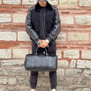 May include: Black leather duffel bag with a reptile skin pattern, featuring two top handles and a shoulder strap. The bag is held by a person wearing a matching black leather baseball cap and a black jacket with a fur collar.