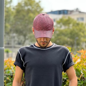Può includere: Un cappellino da baseball bordeaux con un motivo strutturato simile alla pelle di serpente. Il cappellino ha una visiera curva e un colore bordeaux abbinato. La persona nell'immagine indossa una maglietta nera con cuciture bianche.