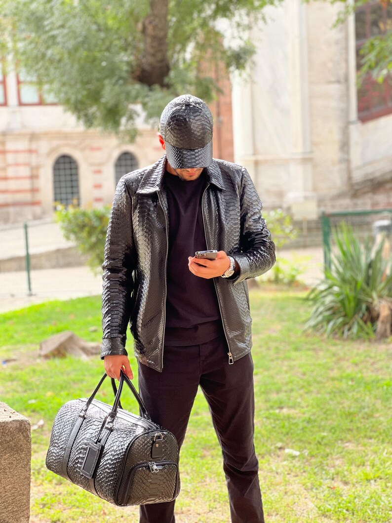 Pode incluir: Casaco de couro preto, bon&eacute; de beisebol e mala de viagem com um padr&atilde;o texturizado. O casaco tem gola e fecho de correr. A mala de viagem tem uma pega superior e um bolso lateral. A pessoa est&aacute; a usar uma t-shirt preta e cal&ccedil;as pretas.