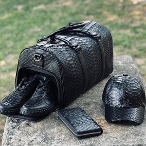 Pode incluir: Uma mala de viagem de couro preta com t&ecirc;nis combinando, uma carteira e um bon&eacute; de beisebol. A mala tem um padr&atilde;o texturizado de pele de cobra. Os t&ecirc;nis est&atilde;o dentro da mala aberta. A carteira e o bon&eacute; est&atilde;o em uma superf&iacute;cie de pedra.