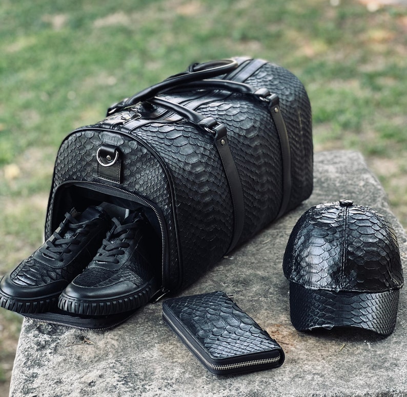 Pode incluir: Uma mala de viagem de couro preta com t&ecirc;nis combinando, uma carteira e um bon&eacute; de beisebol. A mala tem um padr&atilde;o texturizado de pele de cobra. Os t&ecirc;nis est&atilde;o dentro da mala aberta. A carteira e o bon&eacute; est&atilde;o em uma superf&iacute;cie de pedra.