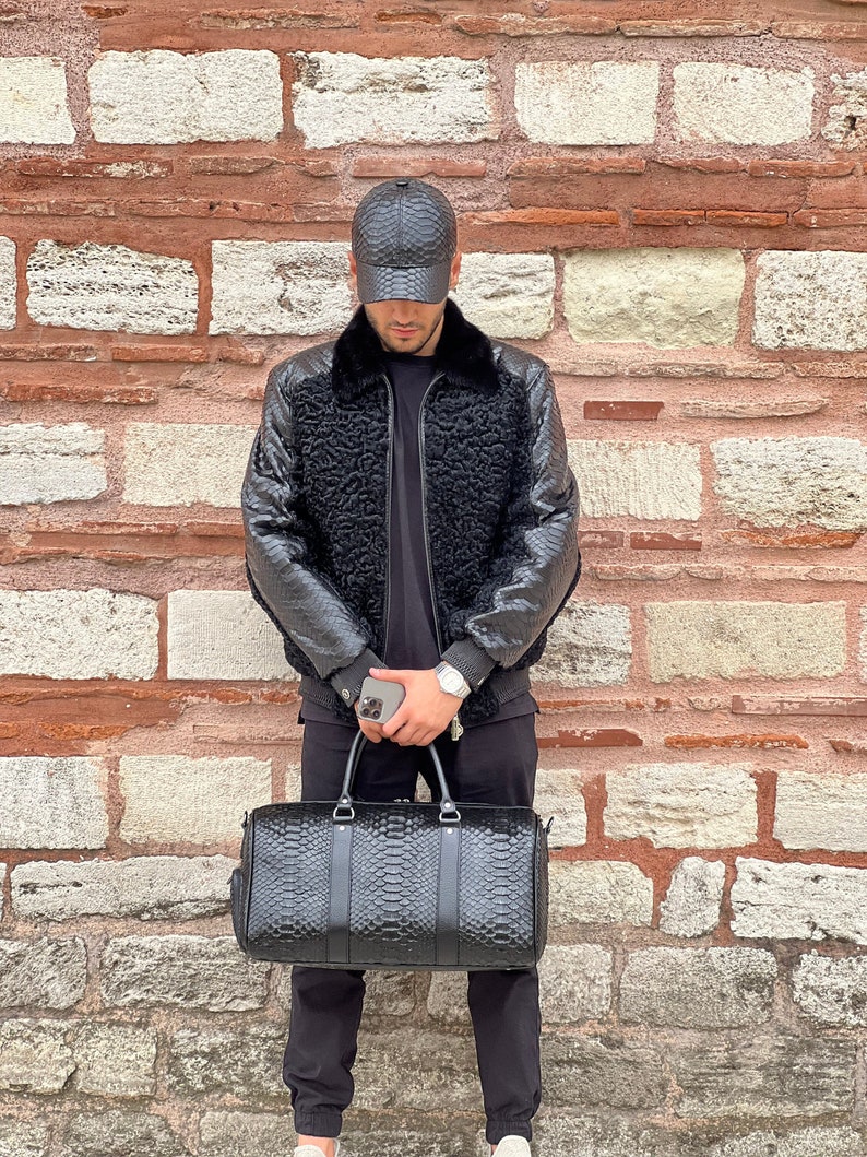 Pode incluir: Mala de viagem de couro preto com padr&atilde;o de pele de r&eacute;ptil, com duas al&ccedil;as superiores e uma al&ccedil;a de ombro. A pessoa est&aacute; usando um bon&eacute; de beisebol de couro preto combinando e uma jaqueta preta com gola de pele.