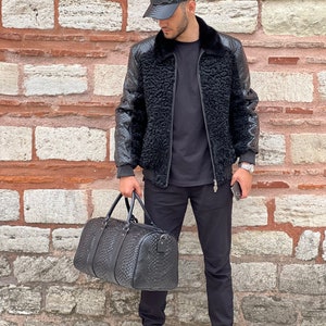 Pode incluir: Casaco bomber de couro preto com gola de pele sint&eacute;tica e bon&eacute; de beisebol a condizer. O casaco tem um padr&atilde;o texturizado e &eacute; combinado com uma mala de viagem preta. O traje &eacute; completado com cal&ccedil;as pretas e t&eacute;nis brancos.
