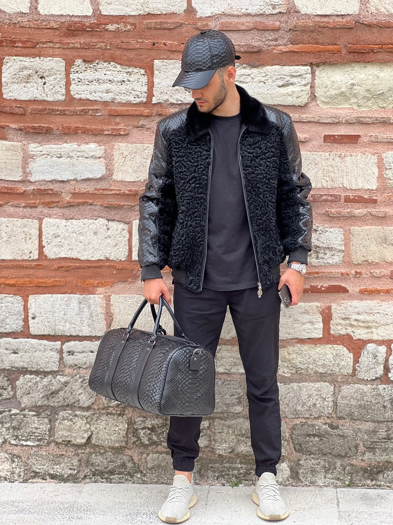 Pode incluir: Casaco bomber de couro preto com gola de pele sint&eacute;tica e bon&eacute; de beisebol a condizer. O casaco tem um padr&atilde;o texturizado e &eacute; combinado com uma mala de viagem preta. O traje &eacute; completado com cal&ccedil;as pretas e t&eacute;nis brancos.