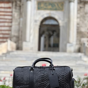 Pode incluir: Uma mala de viagem preta com um padr&atilde;o de pele de cobra, com uma al&ccedil;a superior e uma etiqueta remov&iacute;vel. A mala est&aacute; colocada em frente a um fundo desfocado de um edif&iacute;cio com fachada de pedra e um letreiro verde.