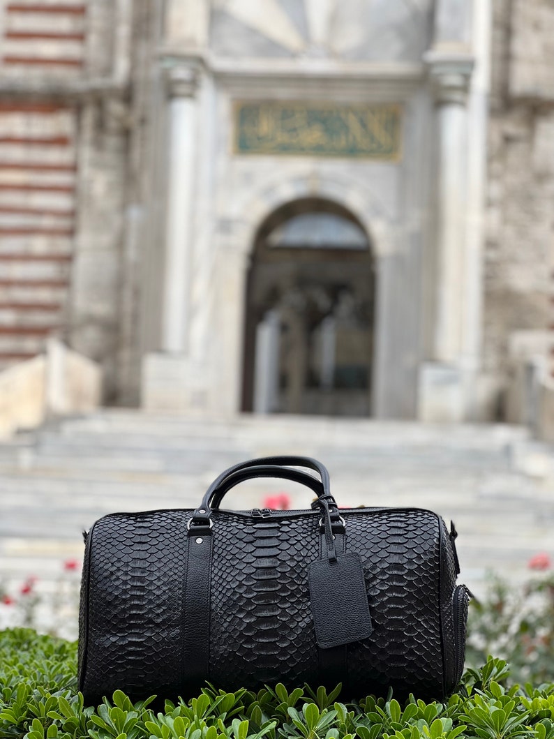 Pode incluir: Uma mala de viagem preta com um padr&atilde;o de pele de cobra, com uma al&ccedil;a superior e uma etiqueta remov&iacute;vel. A mala est&aacute; colocada em frente a um fundo desfocado de um edif&iacute;cio com fachada de pedra e um letreiro verde.