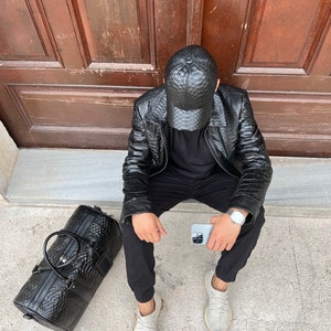 Pode incluir: Casaco e bon&eacute; de beisebol em couro preto texturizado. Uma mala de viagem preta a condizer com uma pega superior e um rel&oacute;gio prateado. A pessoa est&aacute; a usar cal&ccedil;as pretas e t&eacute;nis cinzentos claros. Um smartphone prateado na m&atilde;o.