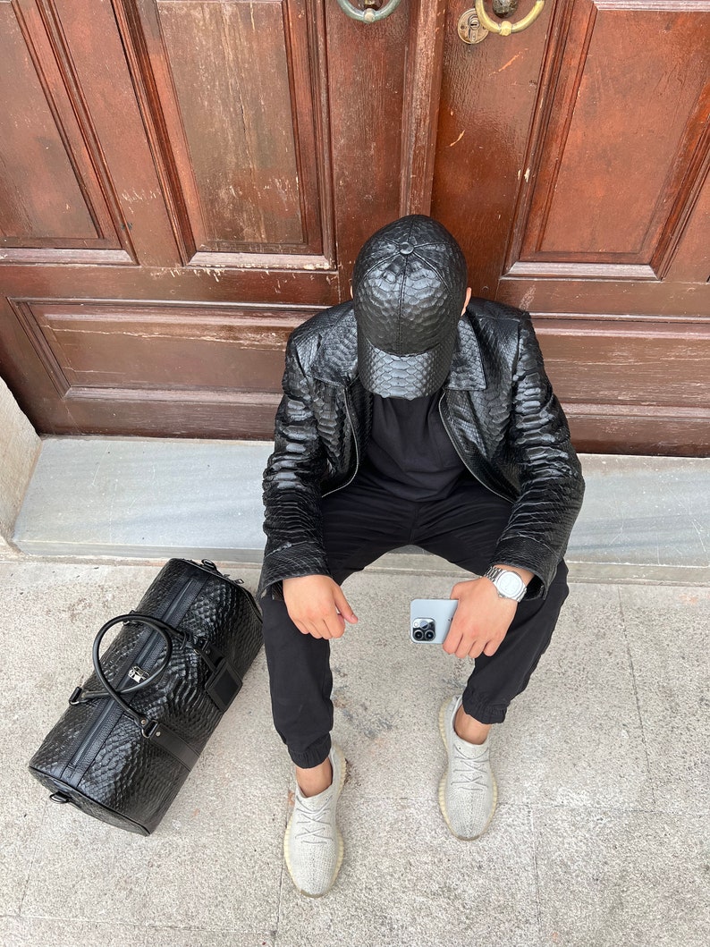 Pode incluir: Casaco e bon&eacute; de beisebol em couro preto texturizado. Uma mala de viagem preta a condizer com uma pega superior e um rel&oacute;gio prateado. A pessoa est&aacute; a usar cal&ccedil;as pretas e t&eacute;nis cinzentos claros. Um smartphone prateado na m&atilde;o.