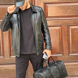 Pode incluir: Casaco, bon&eacute; de beisebol e mala de viagem de couro preto. O casaco tem fecho de correr e gola. O bon&eacute; tem uma aba curva. A mala de viagem tem uma pega superior e uma al&ccedil;a de ombro. Todos os itens t&ecirc;m um padr&atilde;o texturizado semelhante a um r&eacute;ptil.