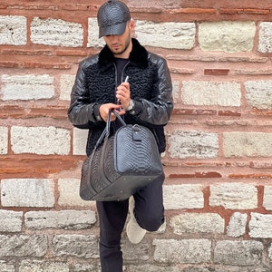 Pode incluir: Uma mala de viagem e bon&eacute; de beisebol de couro preto com um padr&atilde;o texturizado. A mala tem formato arredondado e uma al&ccedil;a superior. O casaco &eacute; preto com gola de pele.